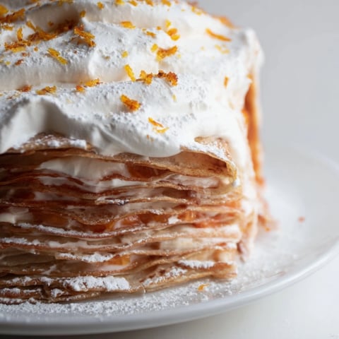 A cross-section of blood orange curd crêpe cake reveals alternating layers of delicate crêpes, vibrant curd, and fluffy whipped cream, garnished with fresh orange slices.
